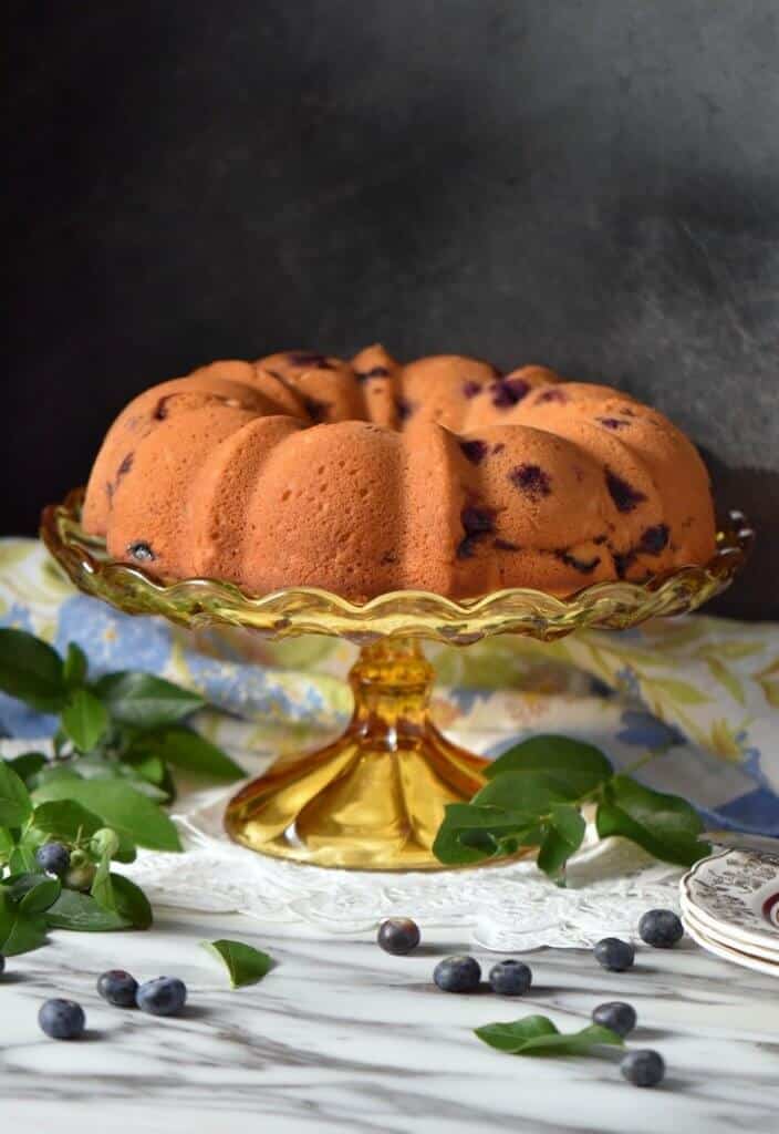 A Limoncello Cake set on an amber, depression glass cake stand, surrounded by fresh blueberries and the leaves of a blueberry bush.