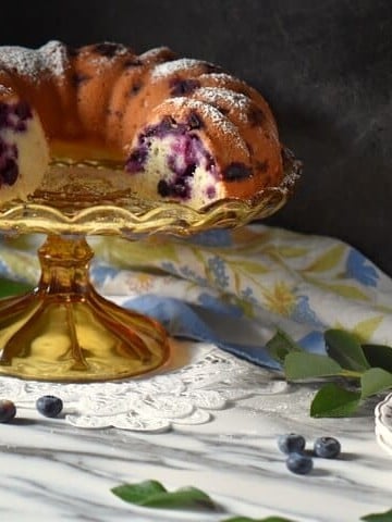 Limoncello cake on a cake stand surrounded by fresh blueberries.
