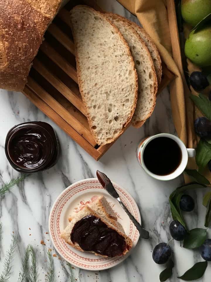 An overhead shot of a loaf of bread,a cup of coffee and plum butter spread thickly on a slice of bread.