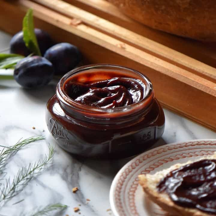 A jar of plum butter is set next to fresh plums.