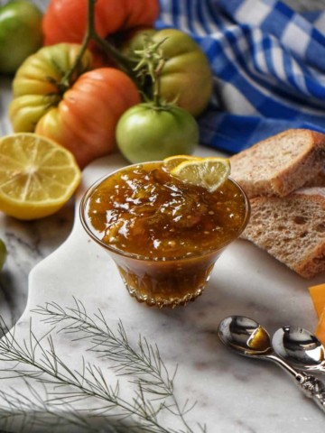 Green tomato jam is in a glass bowl, surrounded by fresh lemons, green tomatoes, and cheese.