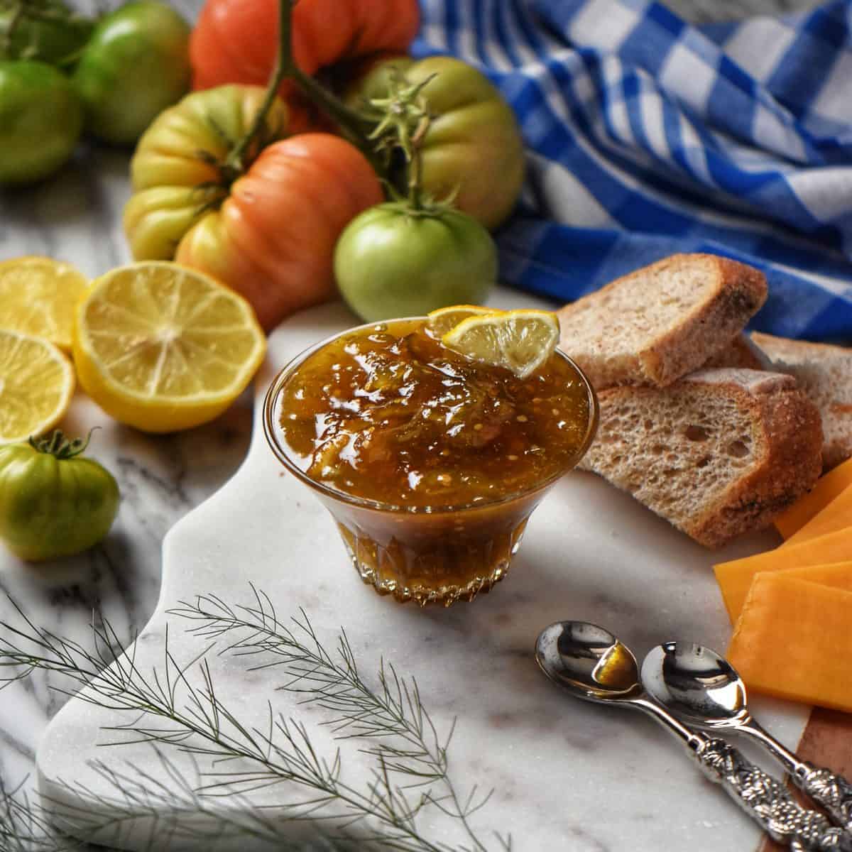 Green tomato jam is in a glass bowl, surrounded by fresh lemons, green tomatoes, and cheese.