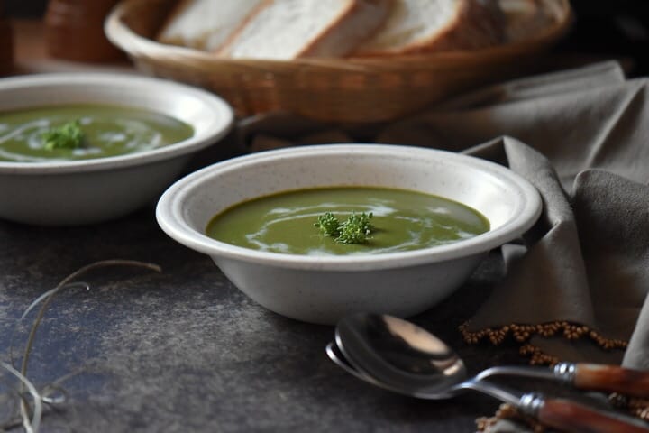Two bowls of Cream of Broccoli soup with a few tablespoons in the foreground.