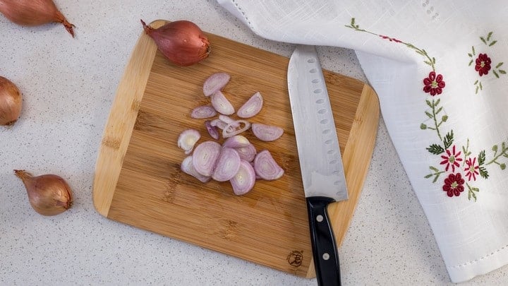 An overhead shot of sliced shallots and a knife on a cutting board; just one of the vegetables that will get oven roasted.