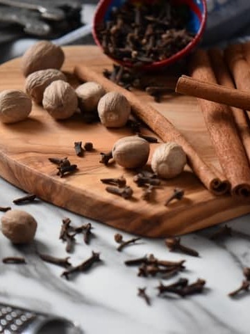 Whole nutmeg, cinnamon sticks and cloves are scattered on a wooden board. Also seen is the result of this allspice recipe, that is to say ground allspice.