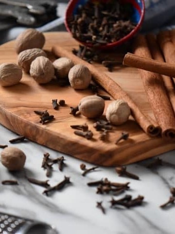 Whole nutmeg, cinnamon sticks and cloves are scattered on a wooden board. Also seen is the result of this allspice recipe, that is to say ground allspice.