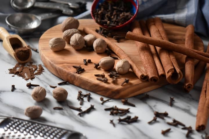 Whole nutmeg, cinnamon sticks and cloves are scattered on a wooden board. Also seen is the result of this allspice recipe, that is to say ground allspice.