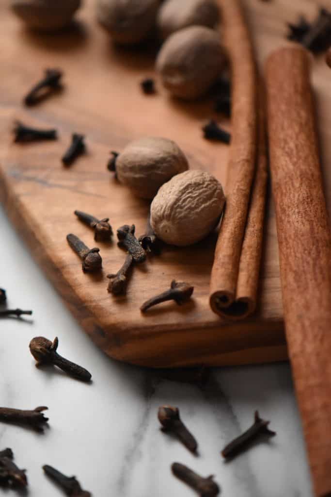 A close up of whole nutmeg, long sticks of cinnamon and cloves on a wooden board.