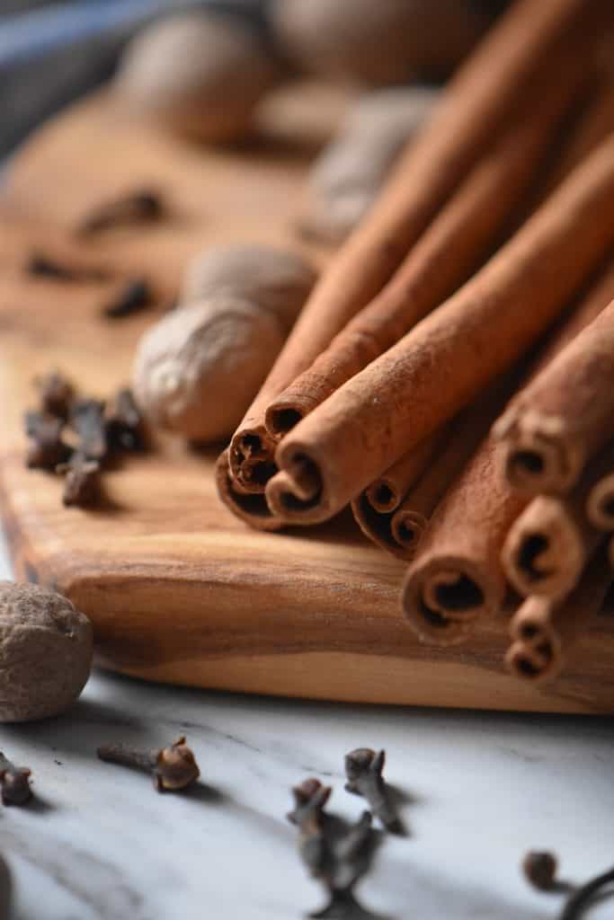 Long cinnamon stick on a wooden board, surrounded by whole nutmeg and cloves; the 3 ingredients required to make an allspice substitute.