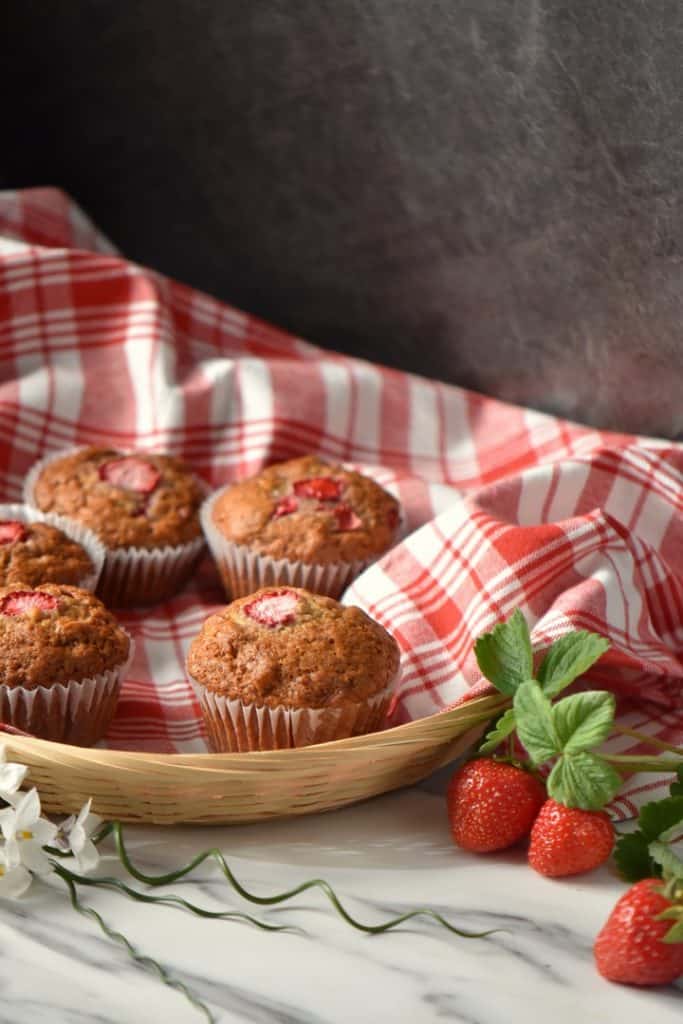 Fresh strawberries surround a wicker basket full of strawberry muffins.