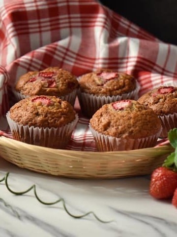 A wicker basket lined with a checkered red tea towel, filled with strawberry muffins.