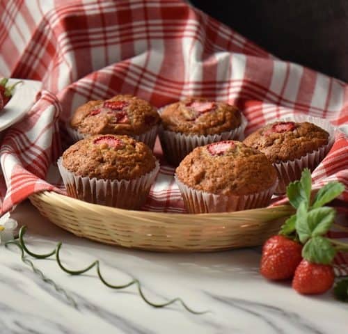 A wicker basket lined with a checkered red tea towel, filled with strawberry muffins.