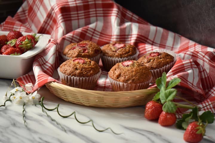 A wicker basket lined with a checkered red tea towel, filled with strawberry muffins.