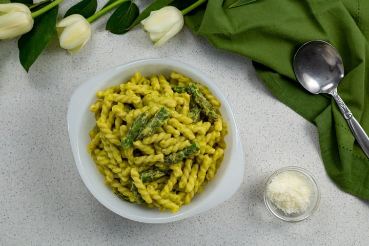 Creamy Asparagus Pasta in a white bowl, next to a small bowl of grated cheese, a green tea towel and tulips.