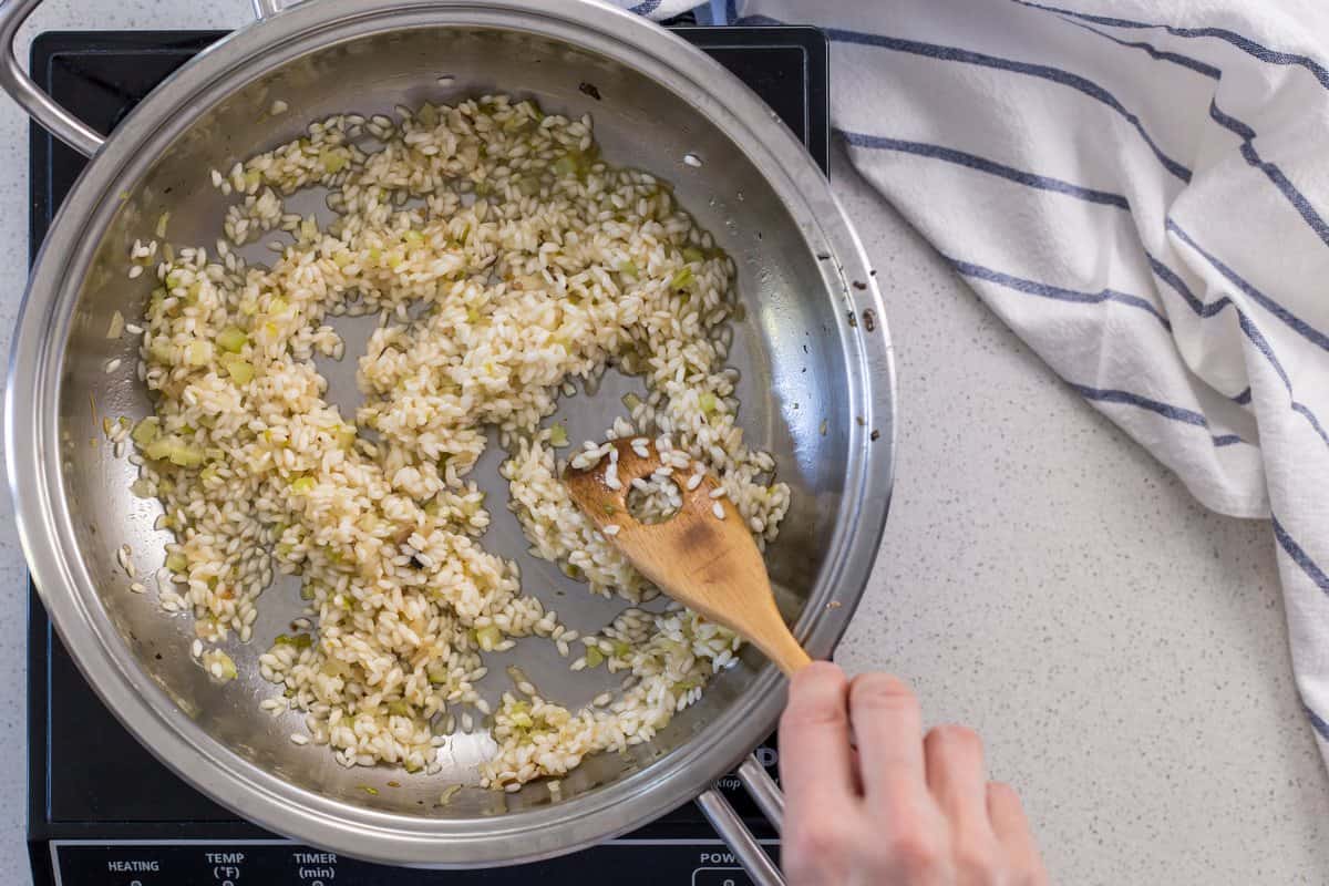 Arborio rice is being mixed with the sauteed shallots and celery in a large pan.