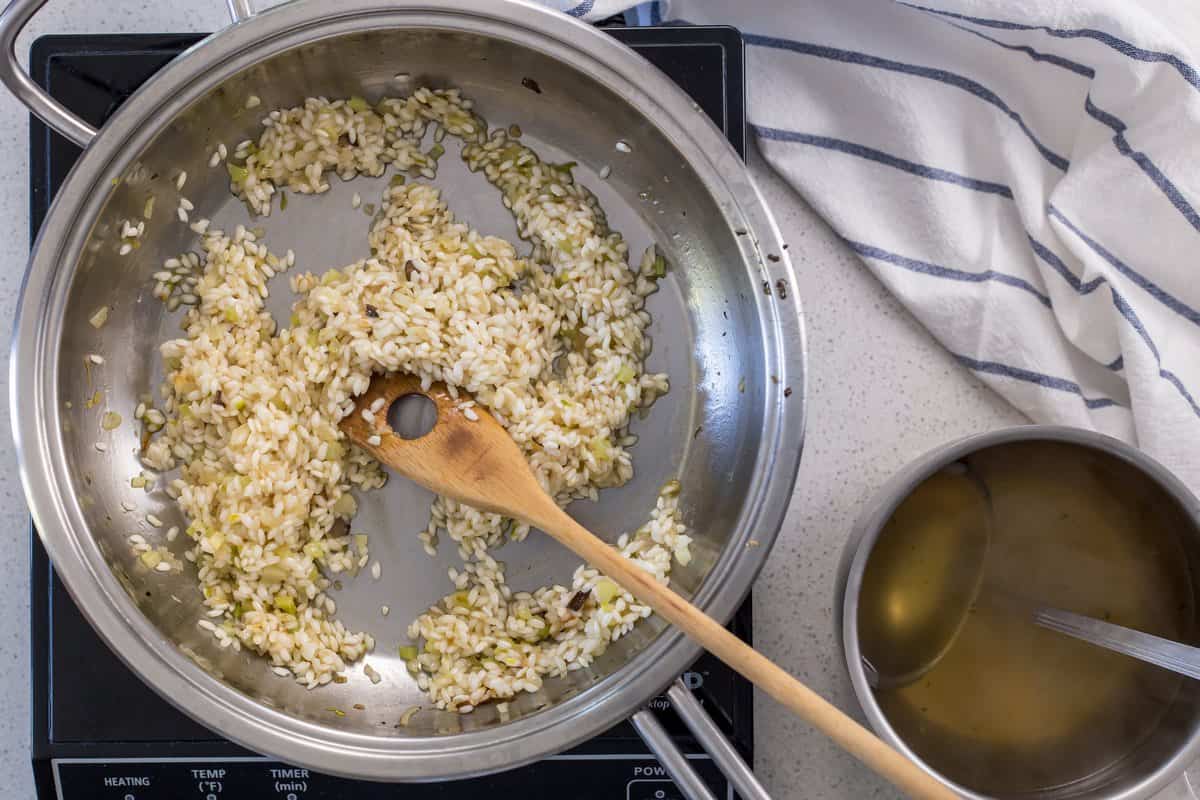 A ladle is in a small pot of broth. It is placed next to a larger pot of arborio rice in need of a ladle of stock. 