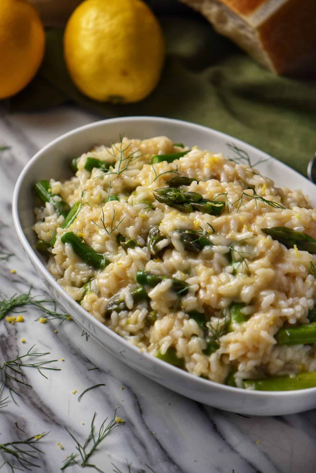 A white oval bowl is filled with creamy asparagus risotto. In the background. a lemon and a block of Parmesan cheese are visible.