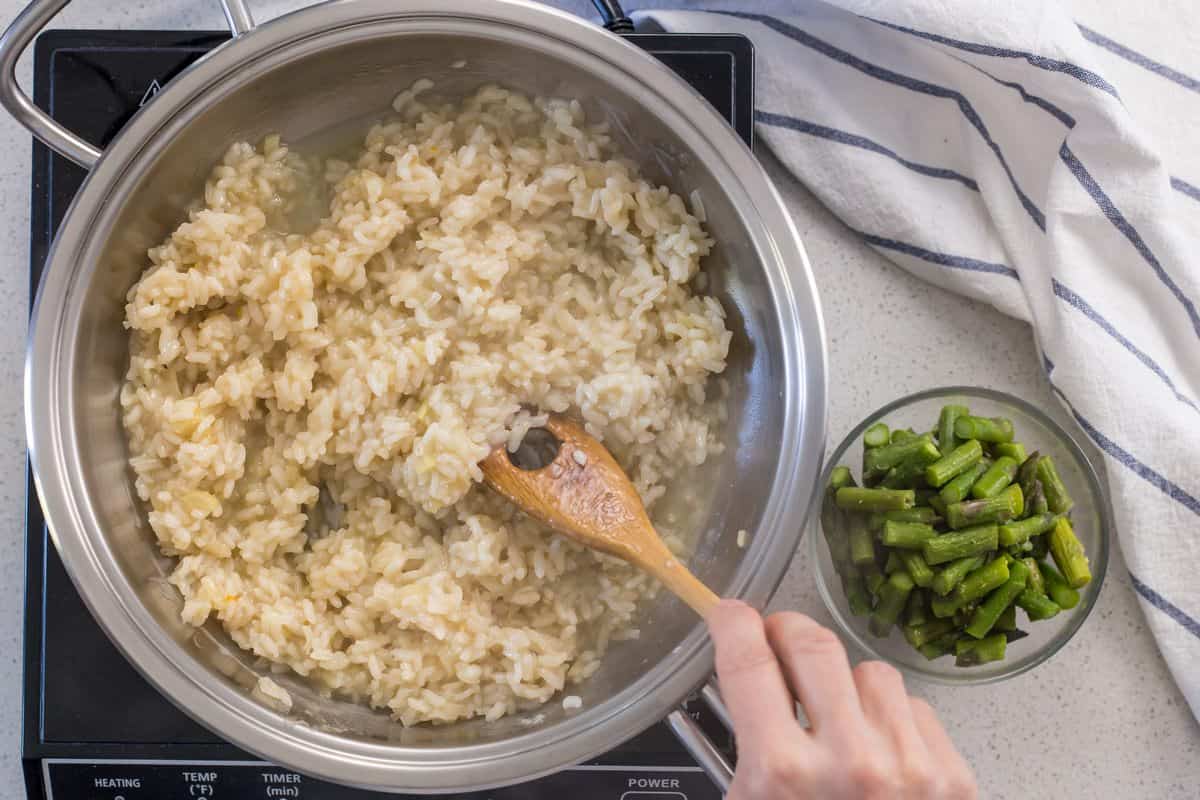 A small dish of cooked chopped asparagus, placed next to a pot of risotto being stirred.