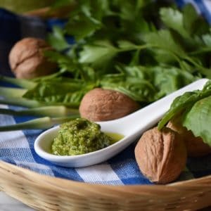 A tablespoon of garlic scape pesto on a checkered tea towel surrounded by fresh basil and parsley.