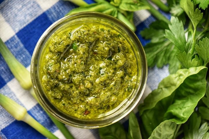 Garlic scape pesto in a mason jar surrounded by basil leaves and a few garlic scapes.