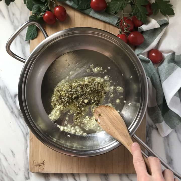 Traditional basil pesto is being added to sauteed garlic in a large pan.