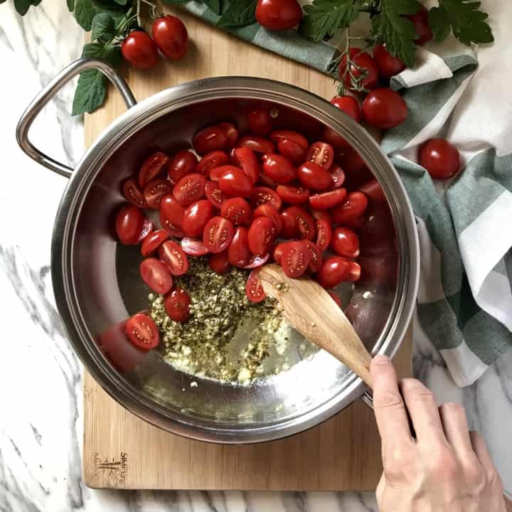 Chopped cherry tomatoes being combined with sauteed garlic and pesto sauce.