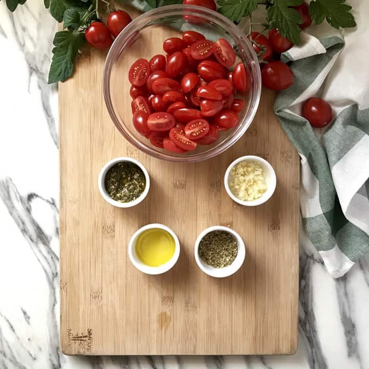 A big bowl of sliced cherry tomatoes next to smaller bowls containing pesto, olive oil, oregano and chopped garlic.
