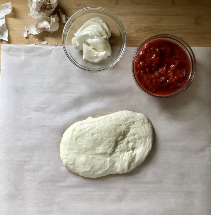 Pizza dough on parchment paper next to a bowl of pizza sauce and mozzarella.