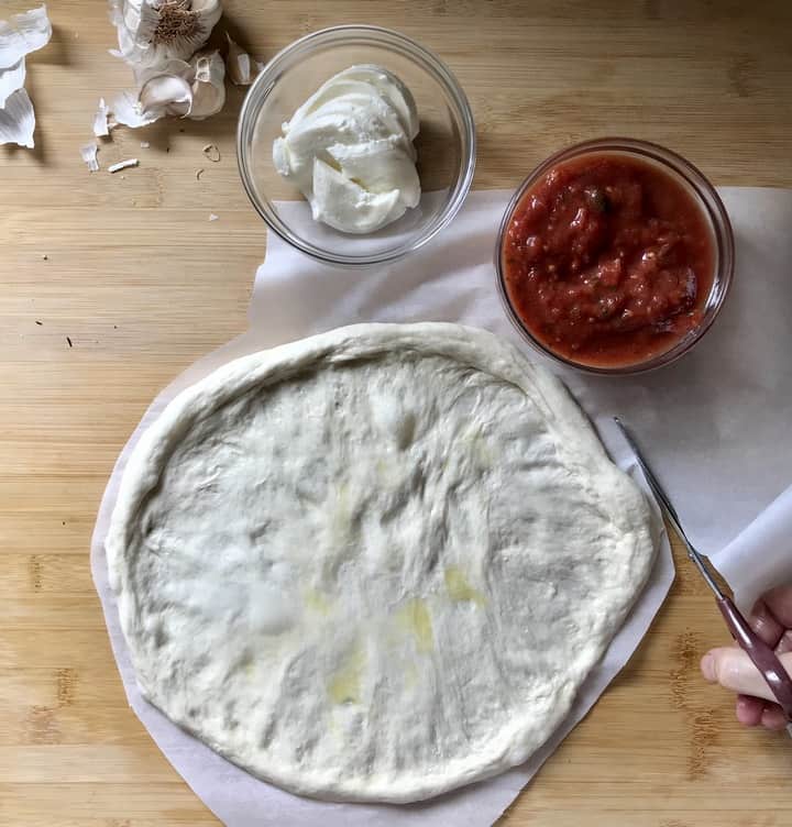 Scissors are being used to trim excess parchment paper around the stretched out pizza.