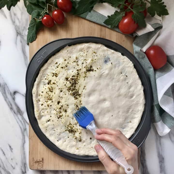 Basil pesto is spread on pizza dough with a pastry brush.