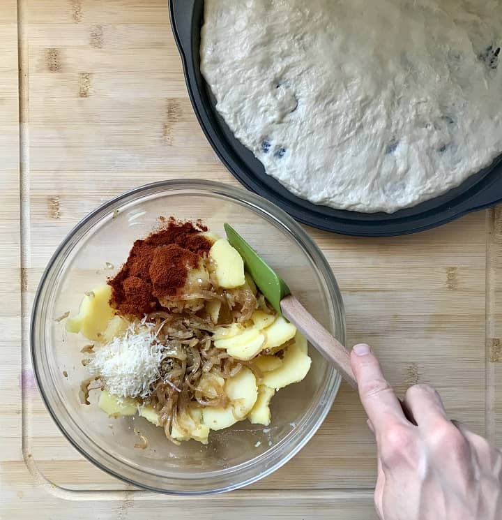 Cooked potatoes being combined with sauteed onions, paprika and grated cheese in a large bowl.