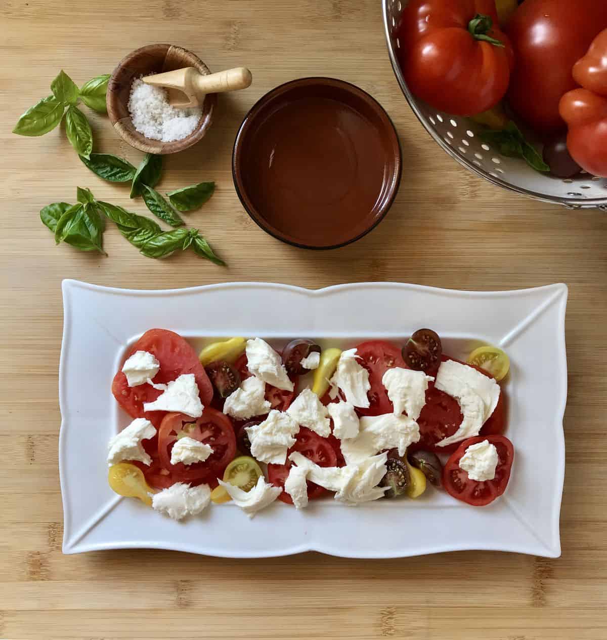 Torn pieces of mozzarella cheese scattered atop sliced heirloom tomatoes in a white serving platter.