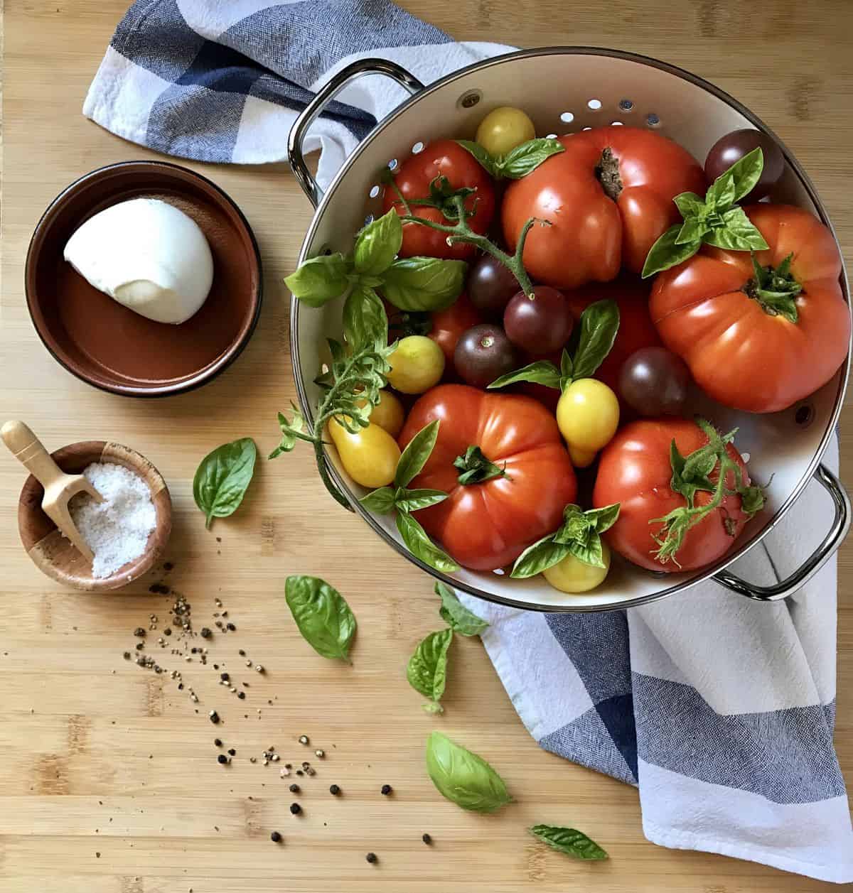A colander filled with heirloom tomatoes, next to basil leaves, a ball of mozzarella, sea salt and pepper.