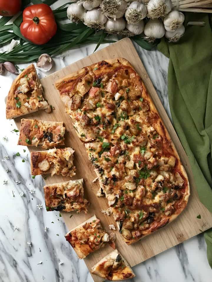 Slices of fish pizza on a wooden board, surrounded by fresh tomatoes and garlic cloves.