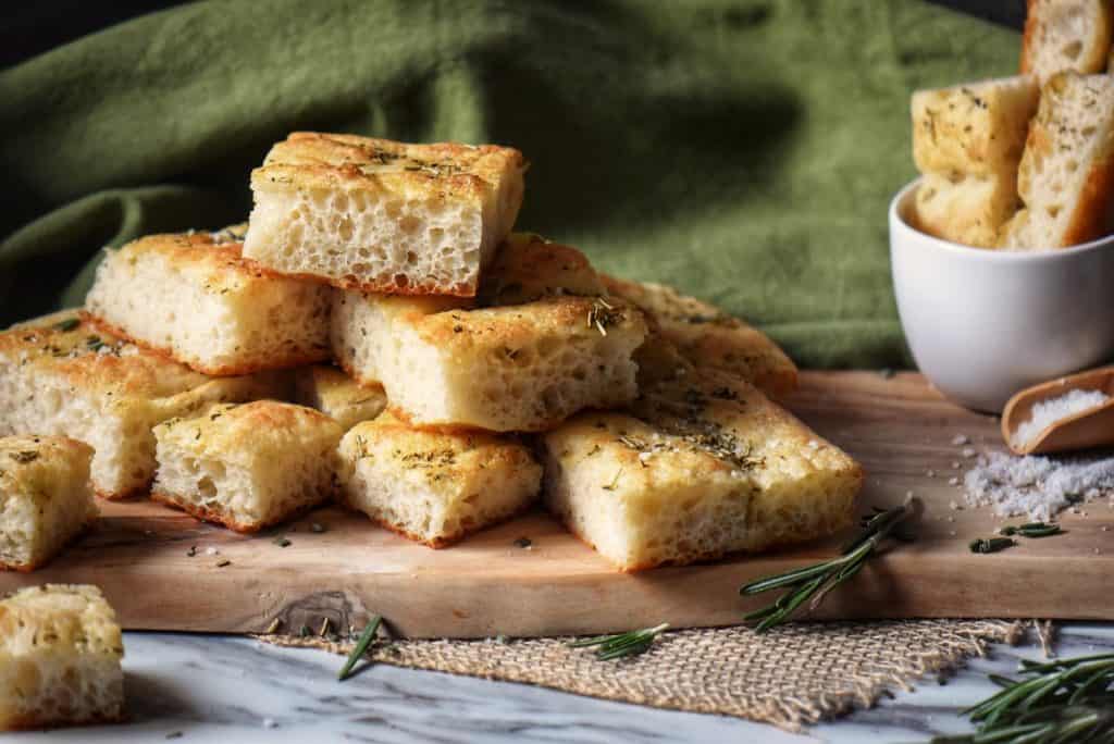 Italian focaccia on a wooden board surrounded by fresh rosemary.