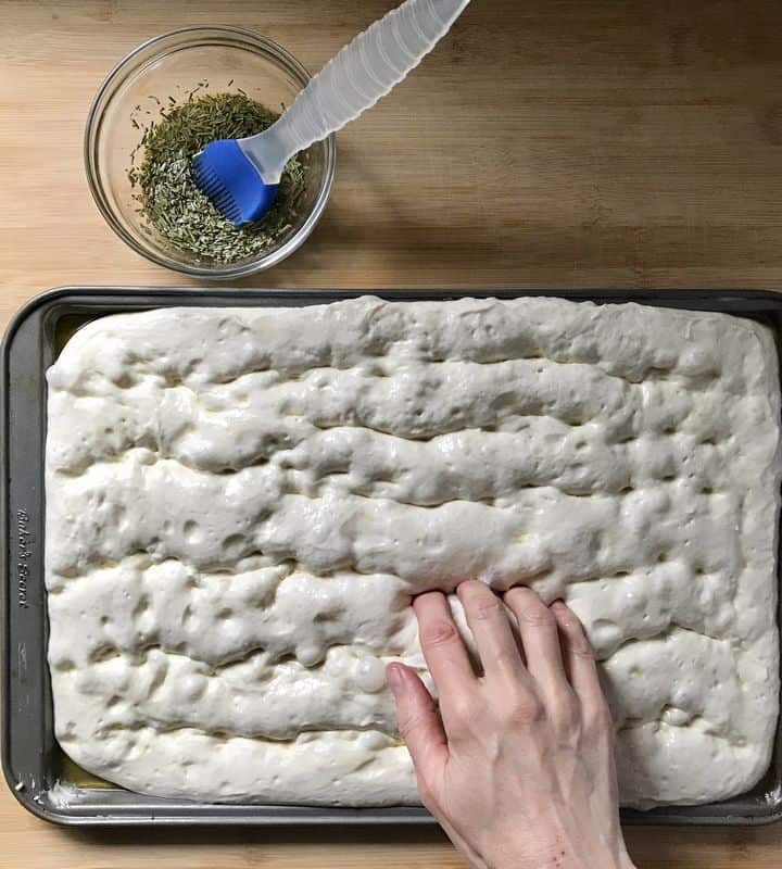 Fingertips are making small dimples in the focaccia dough.