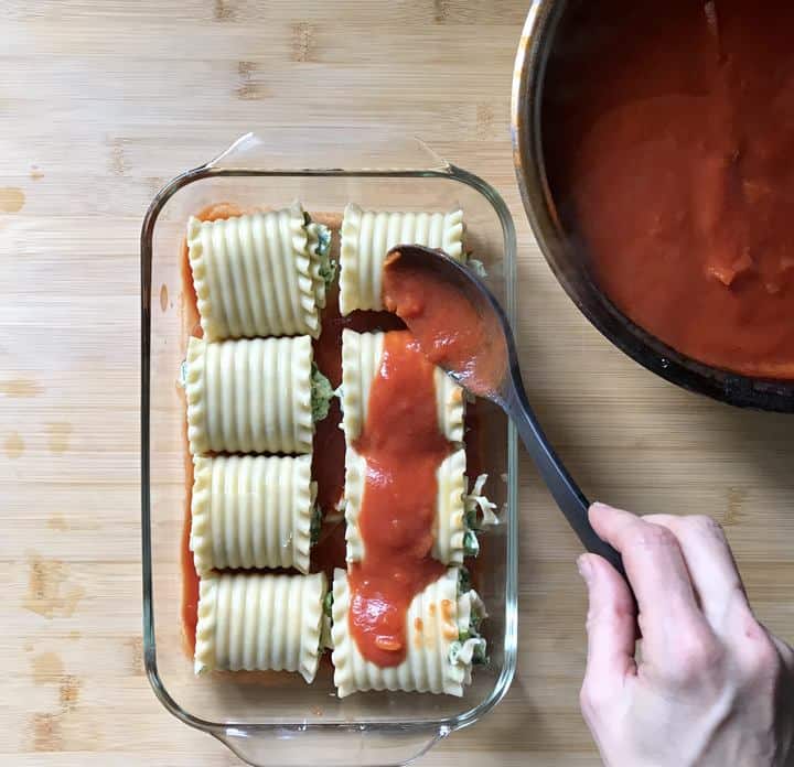 Lasagna roll ups in a baking dish, being topped with tomato sauce.