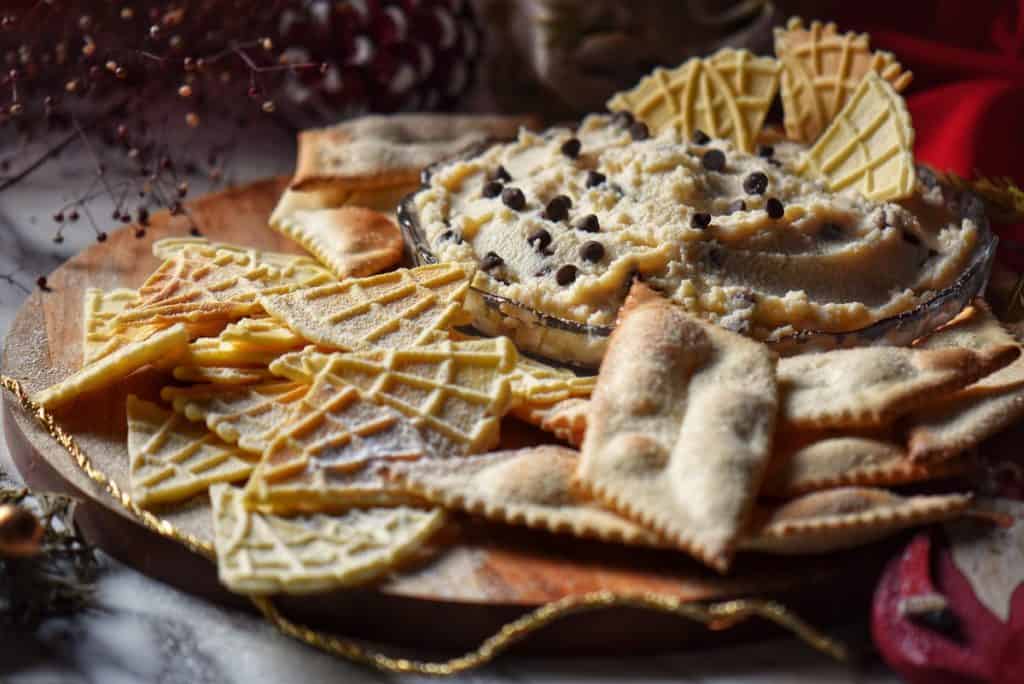 Triangulate pieces of pizzelle next to a bowl of cannoli dip on a wooden board.