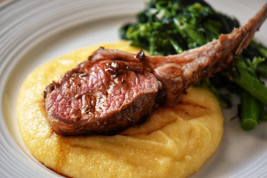 A single lamb rib roast set over creamy polenta in a white dinner plate.