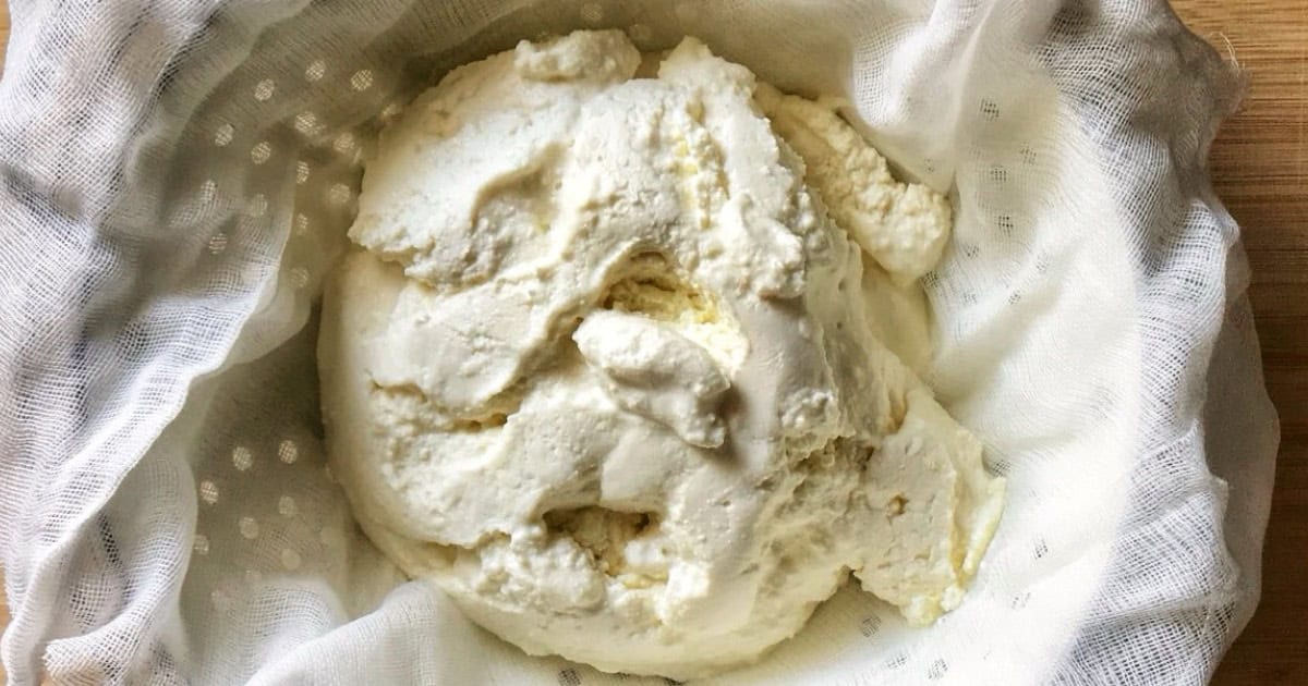 Fresh ricotta cheese being drained in a white cheesecloth-lined strainer over a bowl to remove excess whey.