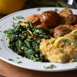 A white dinner plate with cod fish, air fried potatoes and a side dish of sauteed spinach.