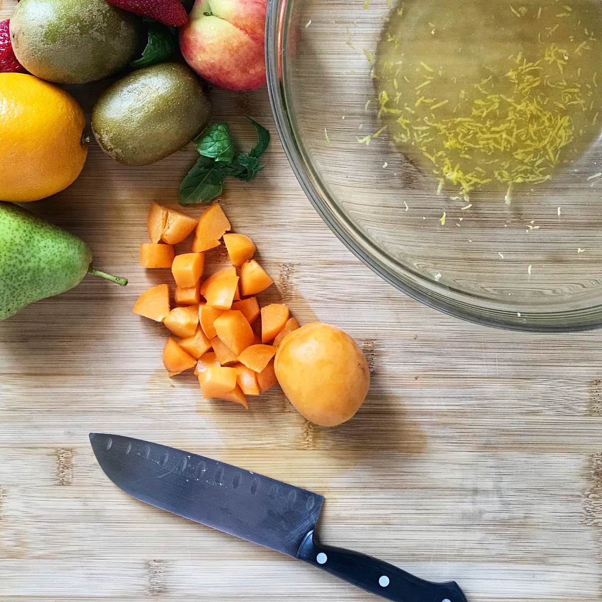 Diced apricots next to fresh fruit and a bowl on a wooden board.