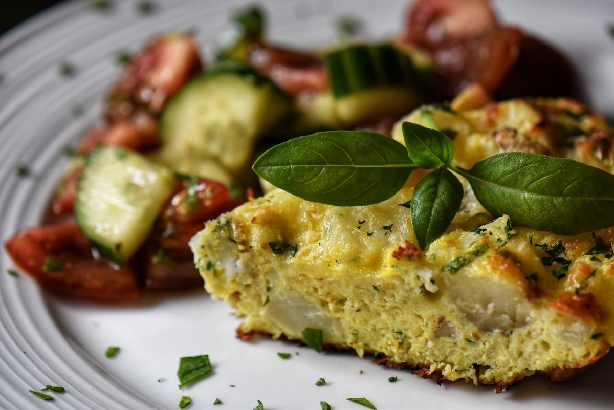 A slice of potato frittata in a white plate served with Italian tomato cucumber salad. 