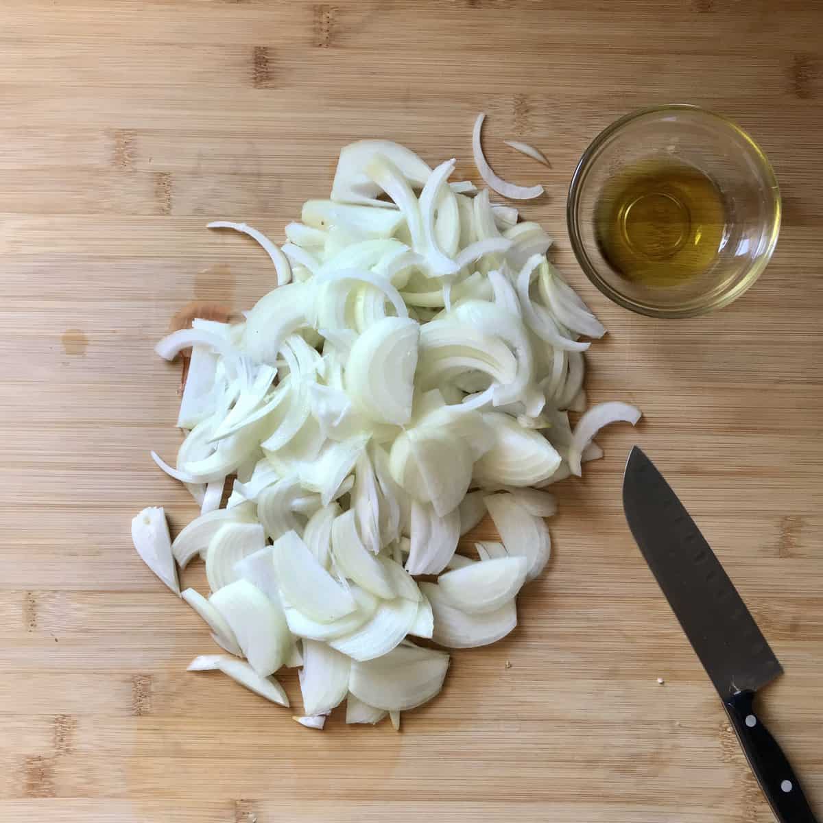Chopped onions on a wooden board next to a knife and a bowl of olive oil.