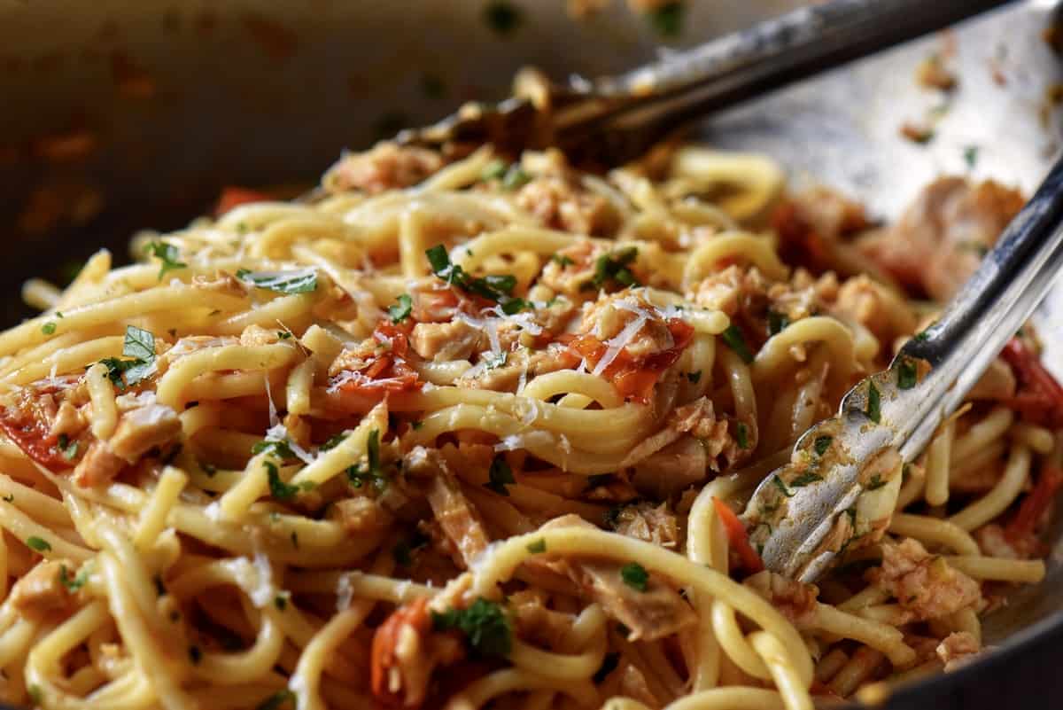 Spaghetti being tossed with cherry tomatoes, tuna, parsley and grated cheese in a pan.