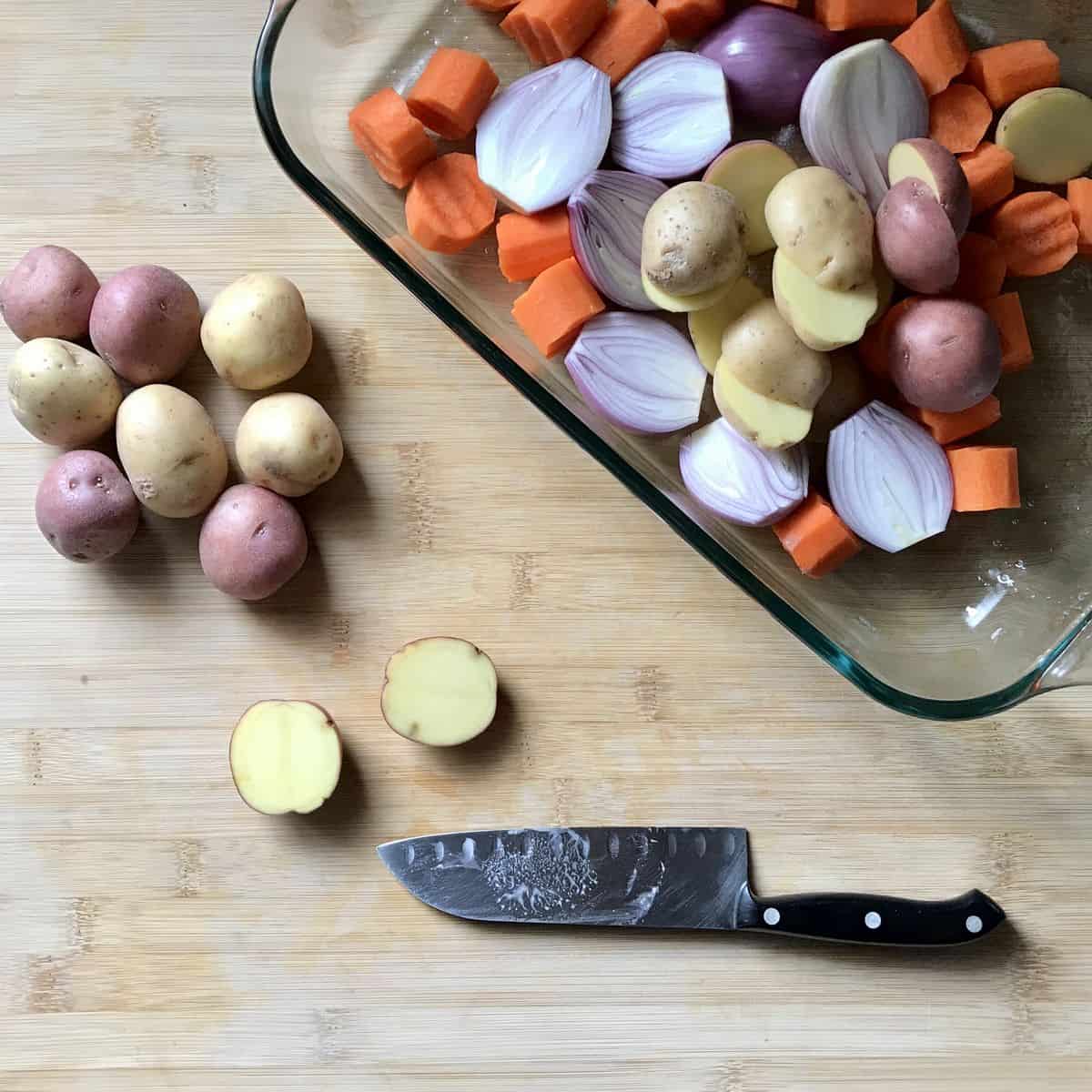 Halved baby potatoes next to a baking dish of sliced carrots and shallots.
