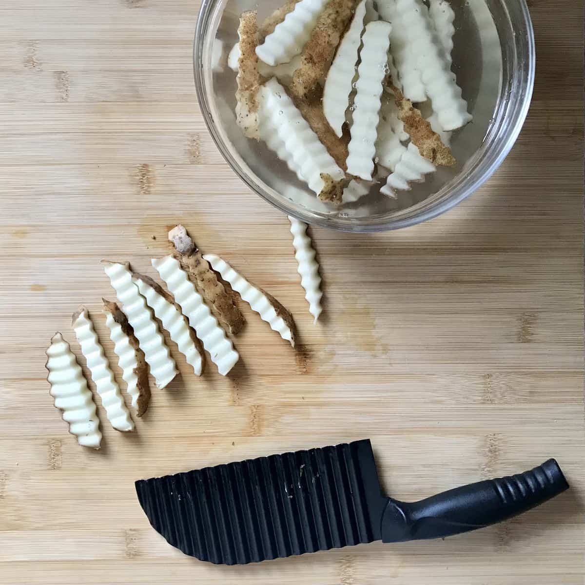 Crinkle cut fries on a wooden board next to a bowl of water with sliced potatoes in it.