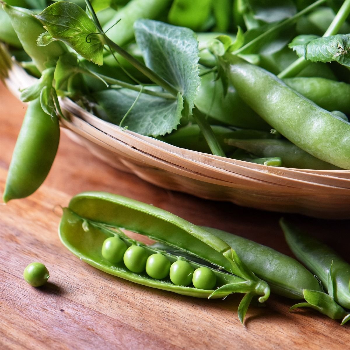 An opened pea shell next to a basket of peas.