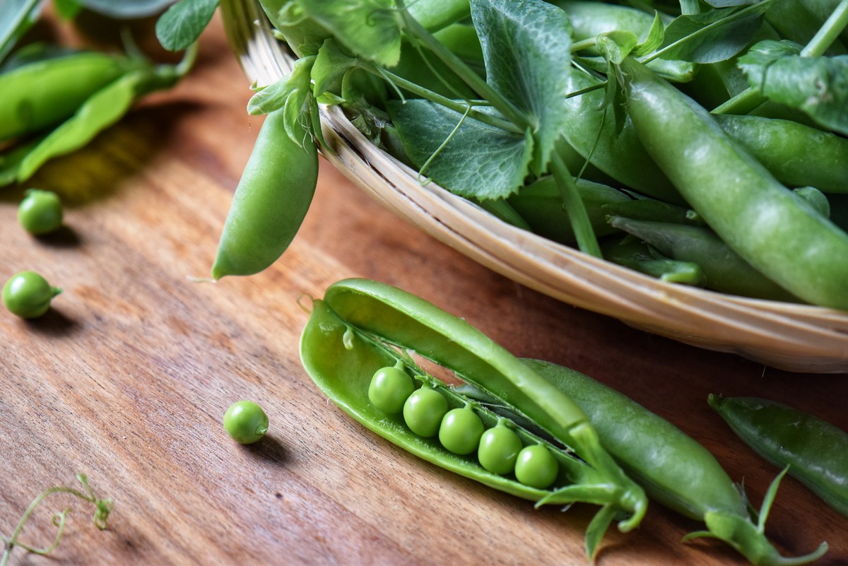 An opened pea shell next to a basket of peas.