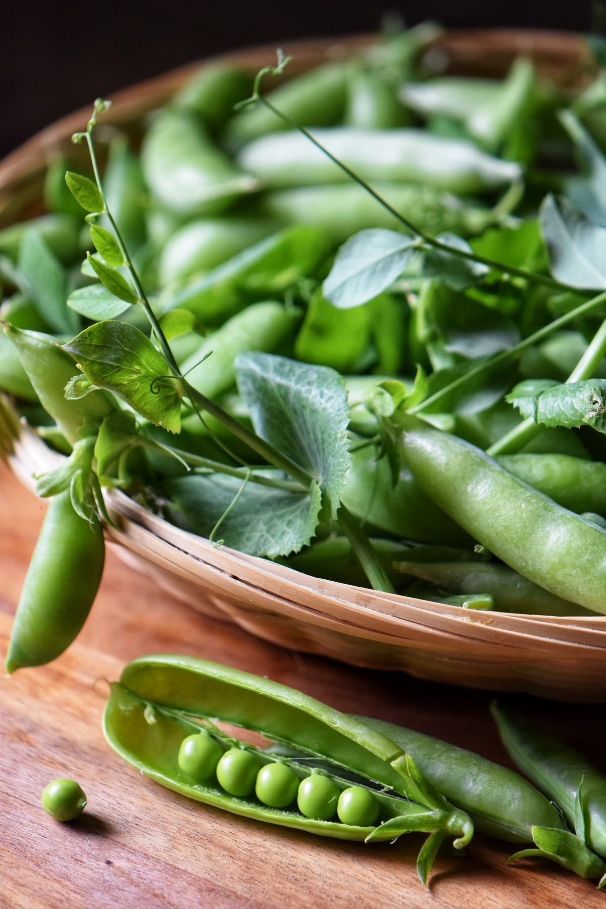 An opened pea shell next to a basket of peas.